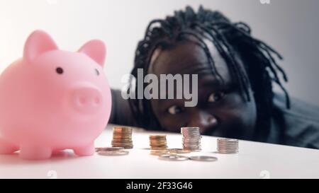 Decisioni finanziarie, investimenti e risparmi. Uomo divertente guardando nelle monete di fronte alla banca piggy. Messa a fuoco della scaffalatura. Foto di alta qualità Foto Stock
