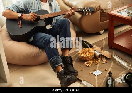 Un giovane creativo seduto a casa sulla borsa dei fagioli con pop corn e bottiglie vuote, reggiseno e denaro sul pavimento, suonando chitarra e canto Foto Stock