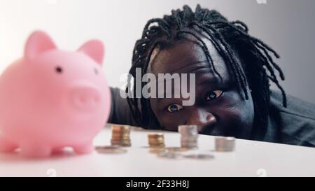 Decisioni finanziarie, investimenti e risparmi. Uomo divertente guardando nelle monete di fronte alla banca piggy. Messa a fuoco della scaffalatura. Foto di alta qualità Foto Stock
