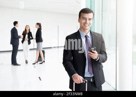 Sorridente uomo d'affari che usa lo smartphone mentre tiene la maniglia del bagaglio in un altro mano al corridoio dell'edificio (terminal dell'aeroporto) Foto Stock