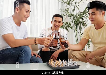 Felici gli amici che brinda con bottiglie di birra mentre si siedono divano e giocare a scacchi a casa Foto Stock