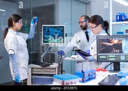 Team di ricercatori medici che conducono lo sviluppo di vaccini con l'aiuto di alta tecnologia, provette, micropipette e scrivere i risultati di analisi su computer in un laboratorio moderno attrezzato di notte Foto Stock