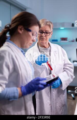 Scienziato professionale e medico senior che discute i risultati degli esami del sangue Tenere un laboratorio dotato di tavoletta Team specialistico di ricercatori medici che lavorano in laboratorio sterile indossare occhiali di protezione Foto Stock