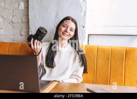 Bruna sorridente giovane donna fotografo che lavora con la sua macchina fotografica e. computer portatile nel caffè Foto Stock