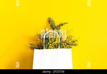 Un bouquet di mimosa in una borsa regalo bianca. Shopping primaverile, regali e promozioni per la Giornata Internazionale della Donna. Sfondo giallo, copyspace. Foto Stock