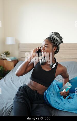 Bella giovane FIT donna nera con bottiglia d'acqua a riposo a letto dopo l'allenamento e parlando al telefono con un amico Foto Stock