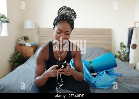 Bella forma giovane donna nera seduta sul letto e la scelta musica per la formazione sul suo smartphone Foto Stock