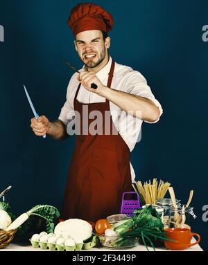 Concetto di pasto vegetariano. Chef con faccia arrabbiata tiene coltelli in metallo Foto Stock