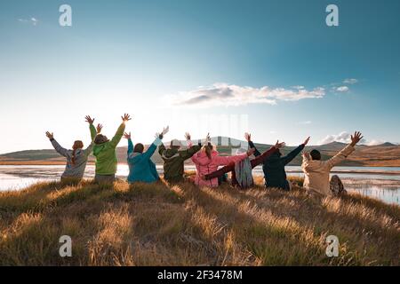 Un grande gruppo di turisti felici si siede con le braccia sollevate al bellissimo tramonto lago e montagne Foto Stock