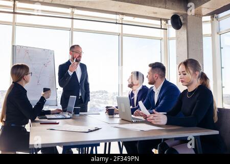 Un uomo d'affari sicuro fa una presentazione di un nuovo progetto nella sala di riunione di un'azienda. Bei revisori parlano con diversi partner abo Foto Stock