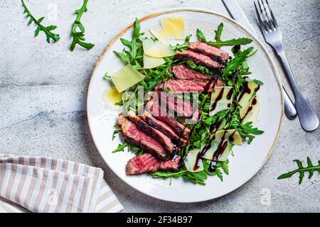 Bistecca di manzo tagliata con rucola e parmigiano su un piatto grigio, fondo grigio. Concetto di cucina Italiana. Foto Stock