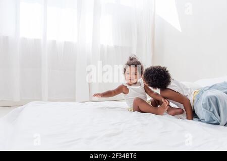 Fratelli e sorelle afroamericani giocano insieme su un letto bianco in un loft interno. Fratelli che si divertono tra i cuscini blu al mattino. Ragazzo baci Foto Stock