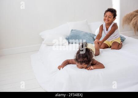 Fratelli e sorelle afroamericani giocano insieme su un letto bianco in un loft interno. Fratelli che si divertono tra i cuscini blu al mattino. Ragazzo pul Foto Stock