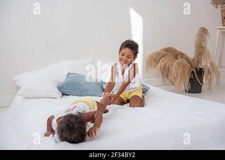 Fratelli e sorelle afroamericani giocano insieme su un letto bianco in un loft interno. Fratelli che si divertono tra i cuscini blu al mattino. Ragazzo pul Foto Stock