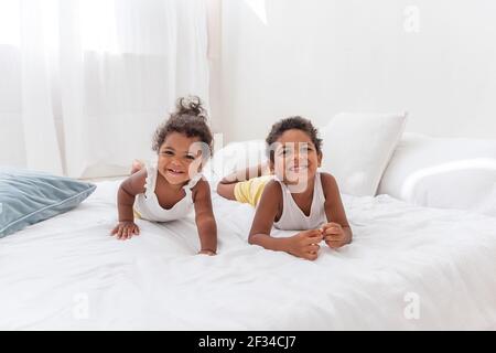 Fratelli e sorelle afroamericani giocano insieme su un letto bianco in un loft interno. Fratelli che si divertono tra i cuscini blu al mattino. Chiudi-u Foto Stock