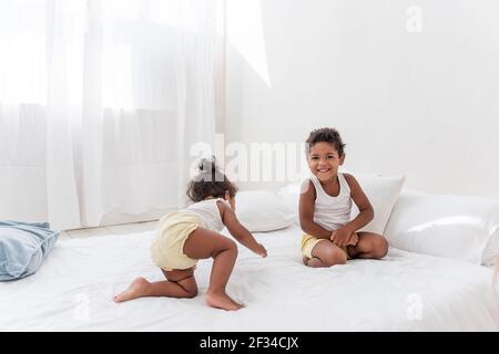 Fratelli e sorelle afroamericani giocano insieme su un letto bianco in un loft interno. Fratelli che si divertono tra i cuscini blu al mattino. Ragazzo pul