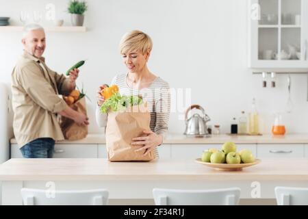 Coppia senior in arrivo dal supermercato con borse della spesa e disimballaggio verdure in cucina al mattino per cucinare Foto Stock