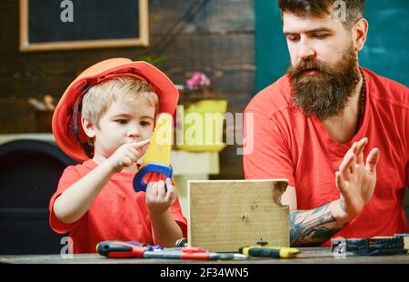 Ragazzo, bambino occupato in casco protettivo imparare ad usare la sega a mano con papà. Concetto di giochi educativi. Padre, genitore con barba che insegna a piccolo figlio Foto Stock