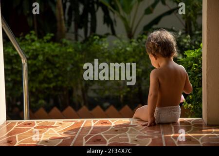 un affascinante bambino si siede sui gradini di una casa su sfondo verde giardino alla luce del sole. vista posteriore Foto Stock