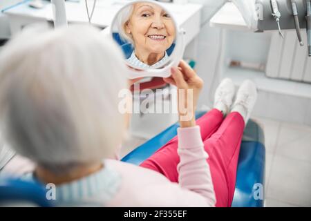 Donna anziano che controlla i suoi denti nello specchio dalla sedia dentale Foto Stock