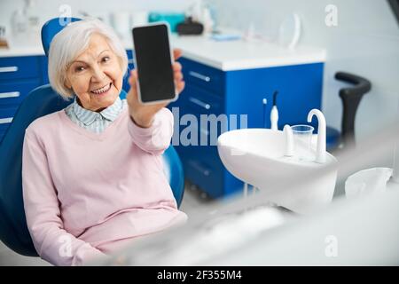 Content pensionato in sedia ospedaliera che dimostra il suo smartphone Foto Stock