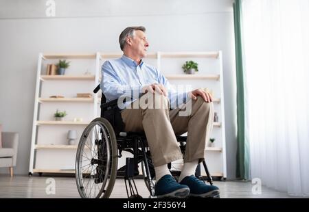 Vista ad angolo basso del paziente di sesso maschile in sedia a rotelle che guarda fuori dalla finestra, sensazione di tristezza e disperazione a casa Foto Stock