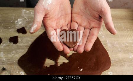 Un giovane chef tiene in mano un cuore fatto di pasta di zenzero, pasta e altre forme sullo sfondo. Concetto di cottura Foto Stock