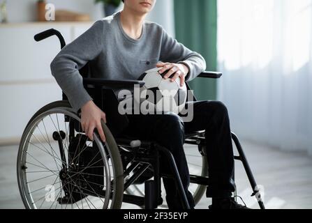 Vista ridotta del ragazzo minorato con la palla di calcio seduta in sedia a rotelle, sentendosi sconvolto sulla sua ferita a casa Foto Stock