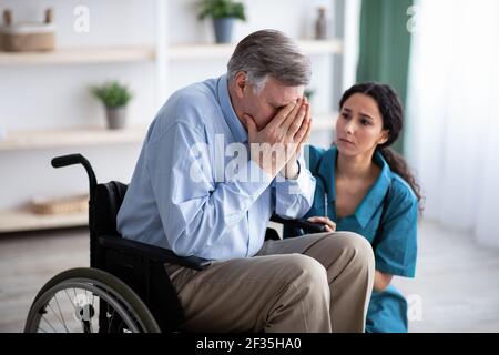 Uomo anziano depresso in sedia a rotelle piangendo, medico giovane cercando di aiutarlo a casa di pensione Foto Stock