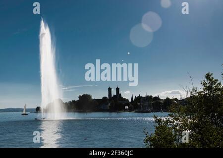 Fontana d'acqua sul lago di Costanza, con un panorama di case sullo sfondo. Foto retroilluminata in tarda estate in una giornata senza nuvole Foto Stock