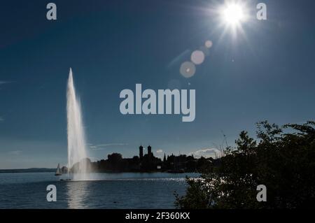 Fontana d'acqua sul lago di Costanza, con un panorama di case sullo sfondo. Foto retroilluminata in tarda estate in una giornata senza nuvole Foto Stock