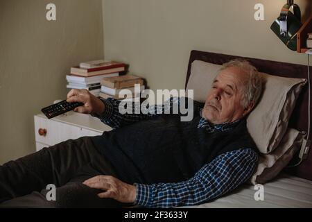Uomo anziano che napping mentre si sdraia nel letto e guardando la TV con un telecomando della televisione, che riposa in un letto Foto Stock