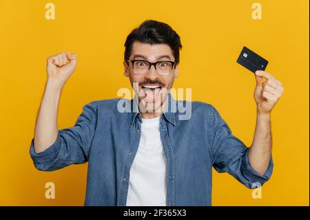 Giovane eccitato positivo uomo caucasico detiene carta di credito, gioisce in successo, vittoria, profitto, in piedi su isolato sfondo arancione alza felicemente il suo pugno e sorridente Foto Stock