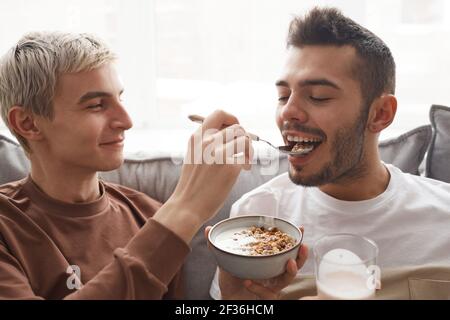 Ritratto di una giocosa coppia gay che gusta la colazione insieme, concentrati su un giovane sorridente che alimenta il ragazzo Foto Stock