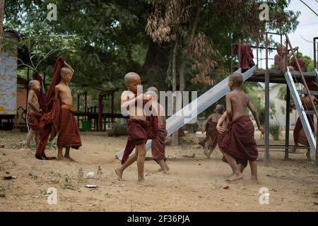 BAGAN, NYAUNG-U, MYANMAR - 2 GENNAIO 2020: Un giovane monaco mangia e mela, mentre gli altri bambini giocano e si divertono in un parco giochi locale con uno scivolo Foto Stock