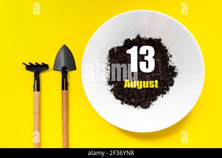 13 agosto. Giorno 13 del mese, data del calendario. Piastra bianca di terreno con una piccola spatola e rastrellare su sfondo giallo. Mese estivo, giorno dell'anno con Foto Stock