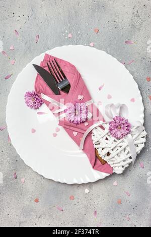 Bel tavolo festivo con piatto bianco, tovagliolo, fiori e decorazioni cuori su sfondo grigio pietra. Vista dall'alto con spazio per la copia. Foto Stock