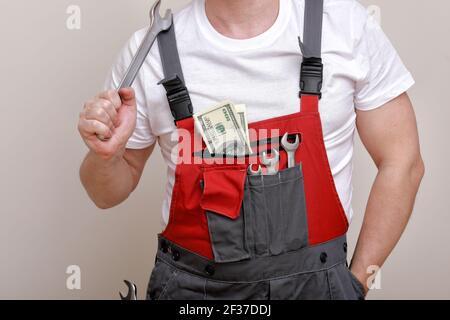foto ritagliata di lavoratore in uniforme rossa, cappello protettivo che tiene dollari, denaro contante e chiave su sfondo bianco. Lavoratore maschile per la pubblicità. Foto Stock