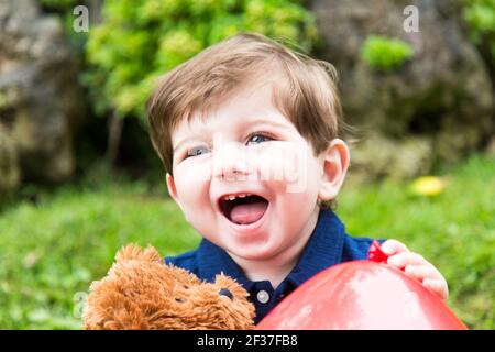 ragazzo piccolo che gioca con un orsacchiotto e un rosso palloncino in giardino Foto Stock