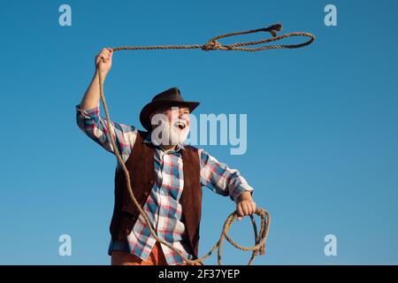 Vecchio cowboy selvaggio West con corda. Uomo occidentale bearded gettando lazo con giacca marrone e cappello che cattura cavallo o mucca. Foto Stock