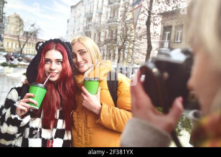 Due carine amiche tengono il caffè e posano per la macchina fotografica mentre la sua amica scatta la foto. Foto Stock