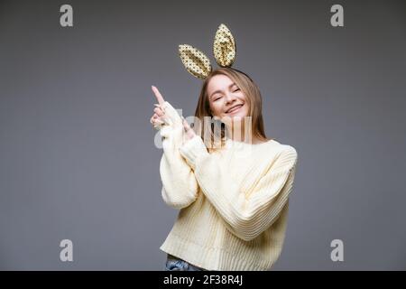 Ritratto di una bella giovane donna allegra in un maglione e nelle orecchie conigliose mostra il suo dito di lato su sfondo grigio Foto Stock