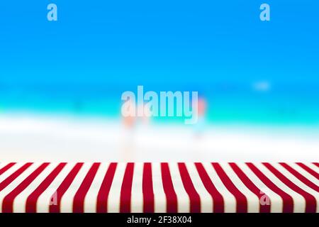 Tavolo coperto con tovaglia rosso striato in sfocatura sfondo spiaggia - può essere utilizzato per la visualizzazione o il montaggio dei prodotti Foto Stock