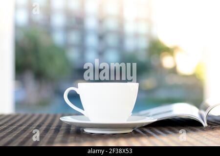 Tazza di caffè sfocata con libro aperto sul tavolo esterno in rattan al mattino, rilassarsi in un resort o in hotel concetto Foto Stock