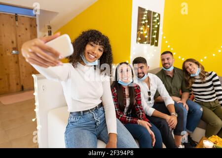 Gruppo di giovani a casa indossando una maschera medica protettiva abbassata in posa per selfie seduta su un divano a casa festa, Afro american ragazza utilizzando sma Foto Stock