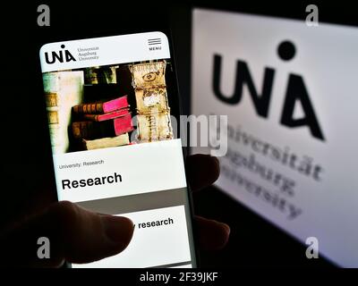 Persona che tiene il telefono cellulare con la pagina web dell'università tedesca Universität Augusta sullo schermo davanti al logo. Mettere a fuoco il centro del display del telefono. Foto Stock