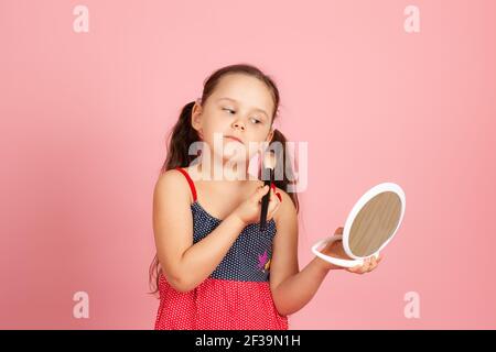 Una ragazza europea con i ponytail e un vestito, che la spolce con un pennello, guarda in uno specchio bianco di plastica, isolato su uno sfondo rosa Foto Stock