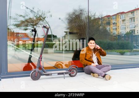 ragazzo che parla giù il cellulare seduto e appoggiato un bicchiere con lo scooter elettrico accanto a lui Foto Stock