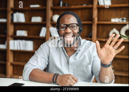 La distanza non è un problema. Positivo nero amichevole uomo in cuffia pratica lingua straniera comunicare online con madrelingua utilizzando il laptop, remoto studente onda mano saluto prof. da videochiamata Foto Stock