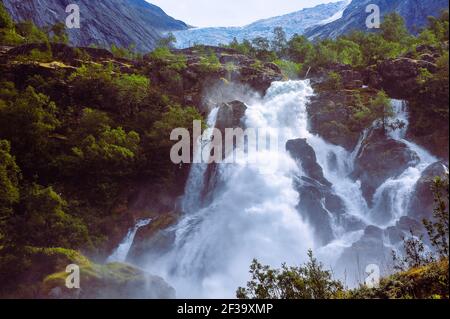 Una cascata vicino al Ghiacciaio Briksdal nel Parco Nazionale del Ghiacciaio Jostedal. Torrent infuriante, acqua bianca, sfondo verde della foresta Foto Stock